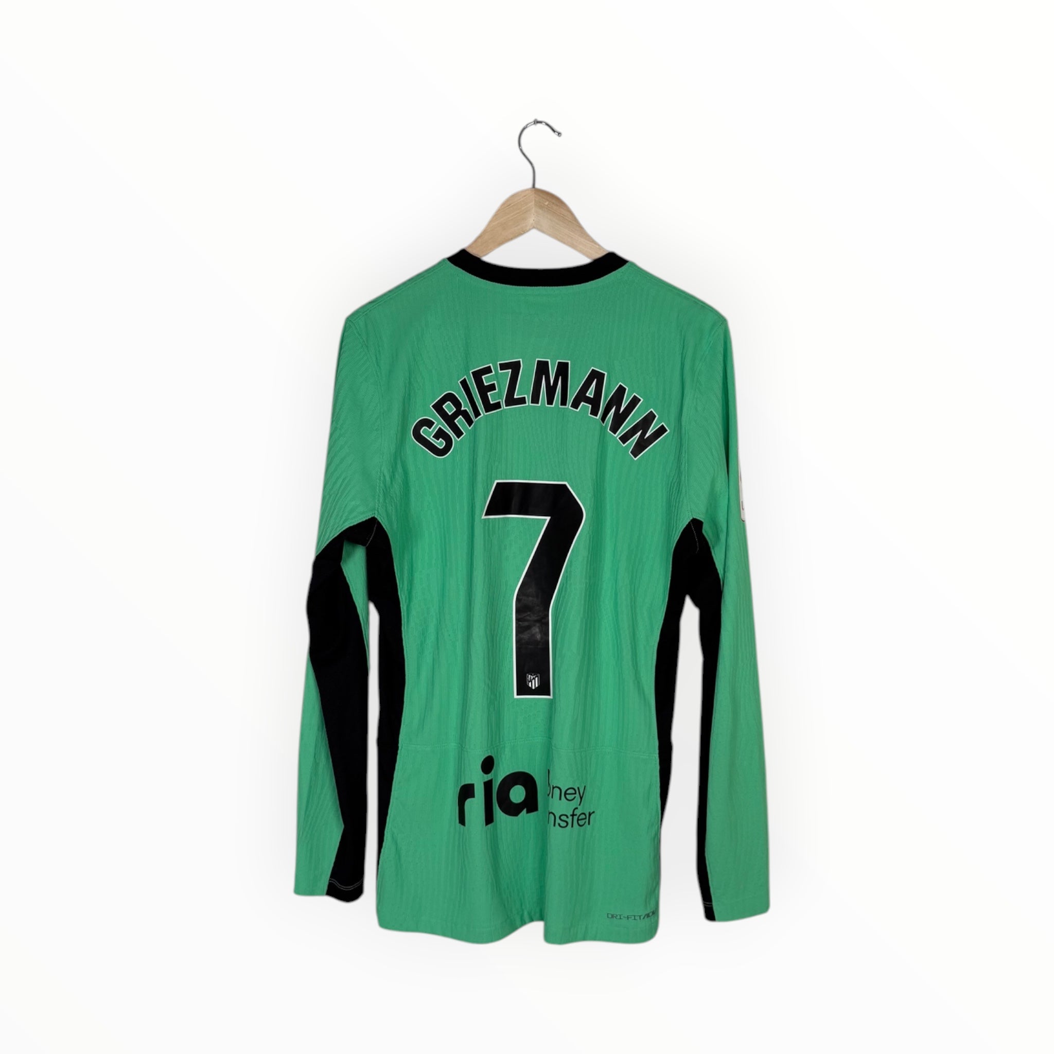 A green long-sleeve soccer jersey with 'GRIEZMANN' and the number '7' printed in black on the back, hanging on a wooden hanger against a plain white background.