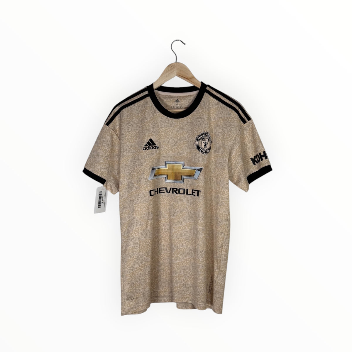 A Manchester United home soccer jersey in a metallic gold-beige colorway, hanging on a wooden hanger against a plain white background. The jersey features black shoulder stripes, black trim on the collar and cuffs, the Adidas logo, the Manchester United crest, the Chevrolet sponsor logo, and a Koho sleeve sponsor logo.