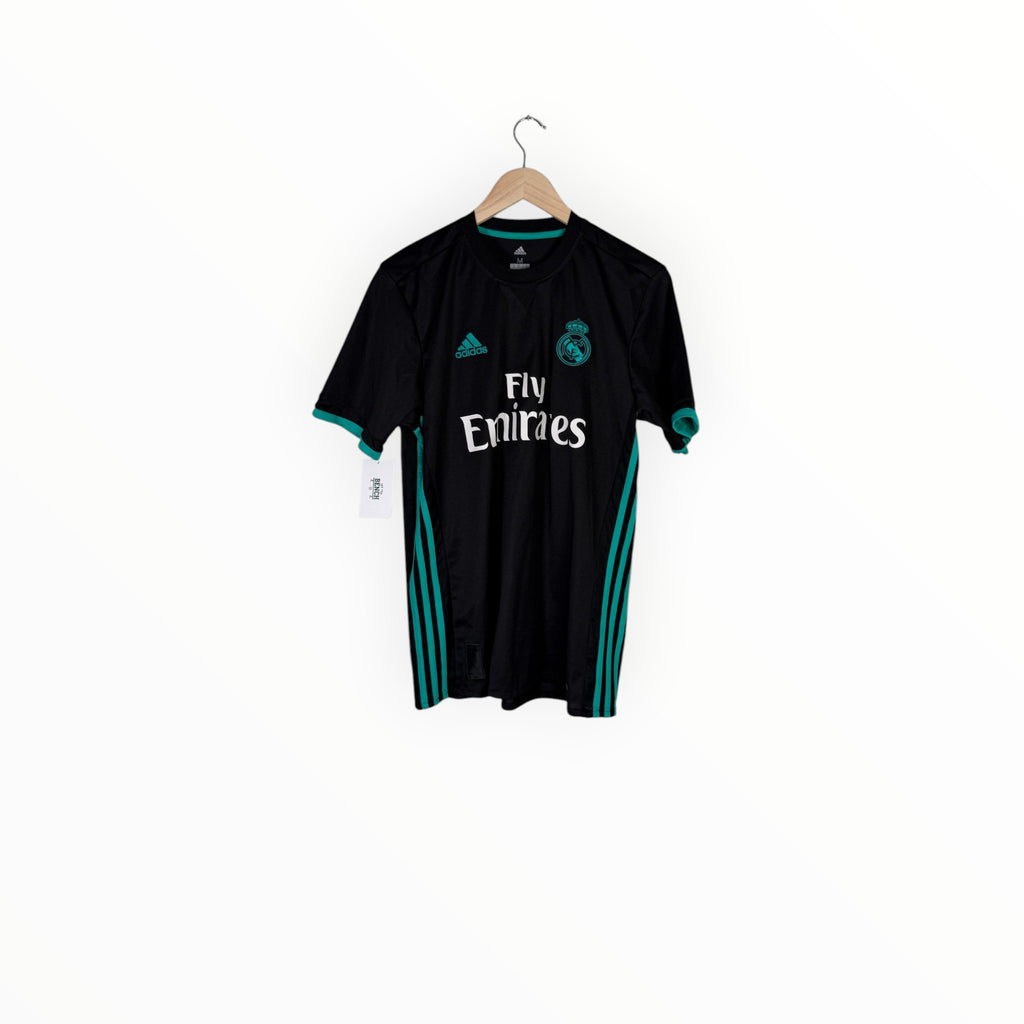 A black Real Madrid soccer jersey with teal accents, hanging on a wooden hanger against a plain white background. The jersey features the white Adidas logo, the Real Madrid crest, and 'Fly Emirates' sponsor text.