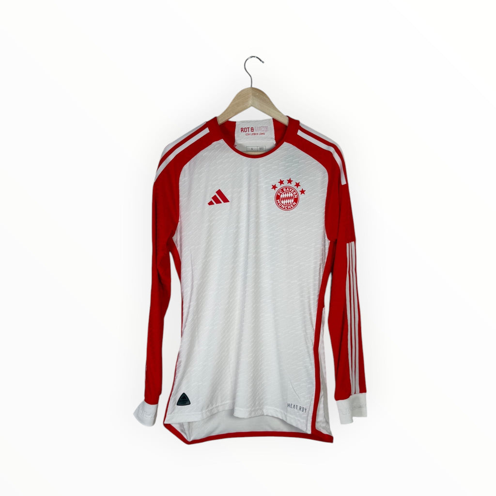A long-sleeve Bayern Munich soccer jersey by Adidas, featuring a white body with red sleeves, red shoulder stripes, and the club's crest on the chest, hanging on a wooden hanger against a white background.