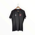 A black Adidas Bayern Munich soccer jersey hanging on a wooden hanger against a plain white wall. The jersey features the Adidas logo on the right chest, the Bayern Munich club crest on the left chest, and three black stripes running down the sides.