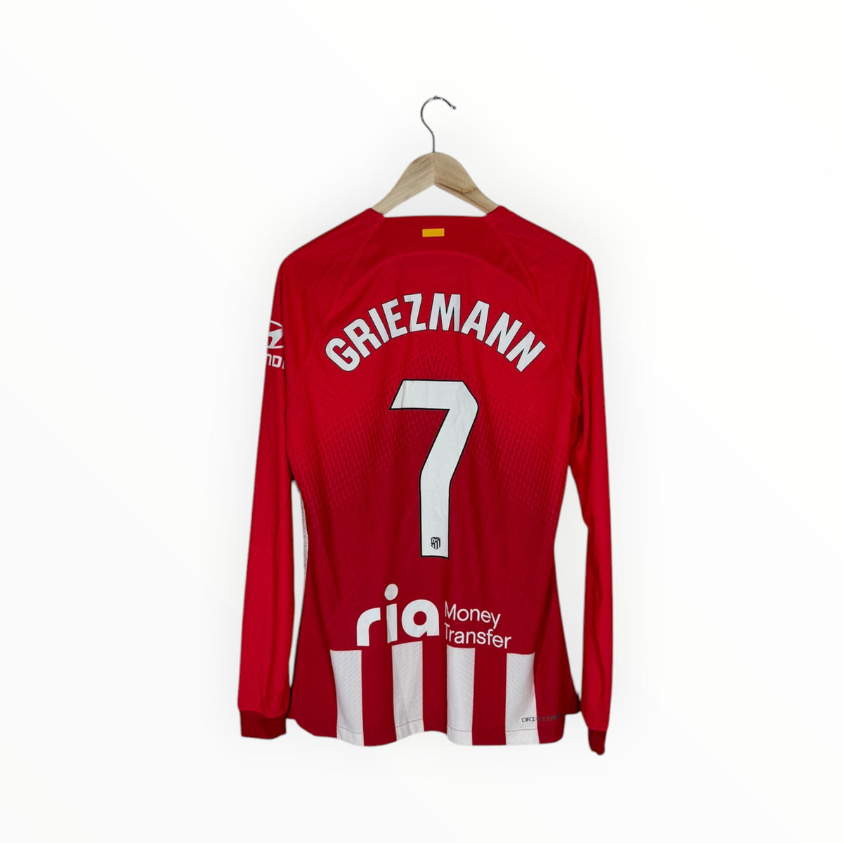 A red long-sleeve soccer jersey with 'GRIEZMANN' and the number '7' printed in white on the back, hanging on a wooden hanger against a plain white wall. The jersey features white vertical stripes near the bottom and a sponsor logo 'rio Money transfer' on the lower back.
