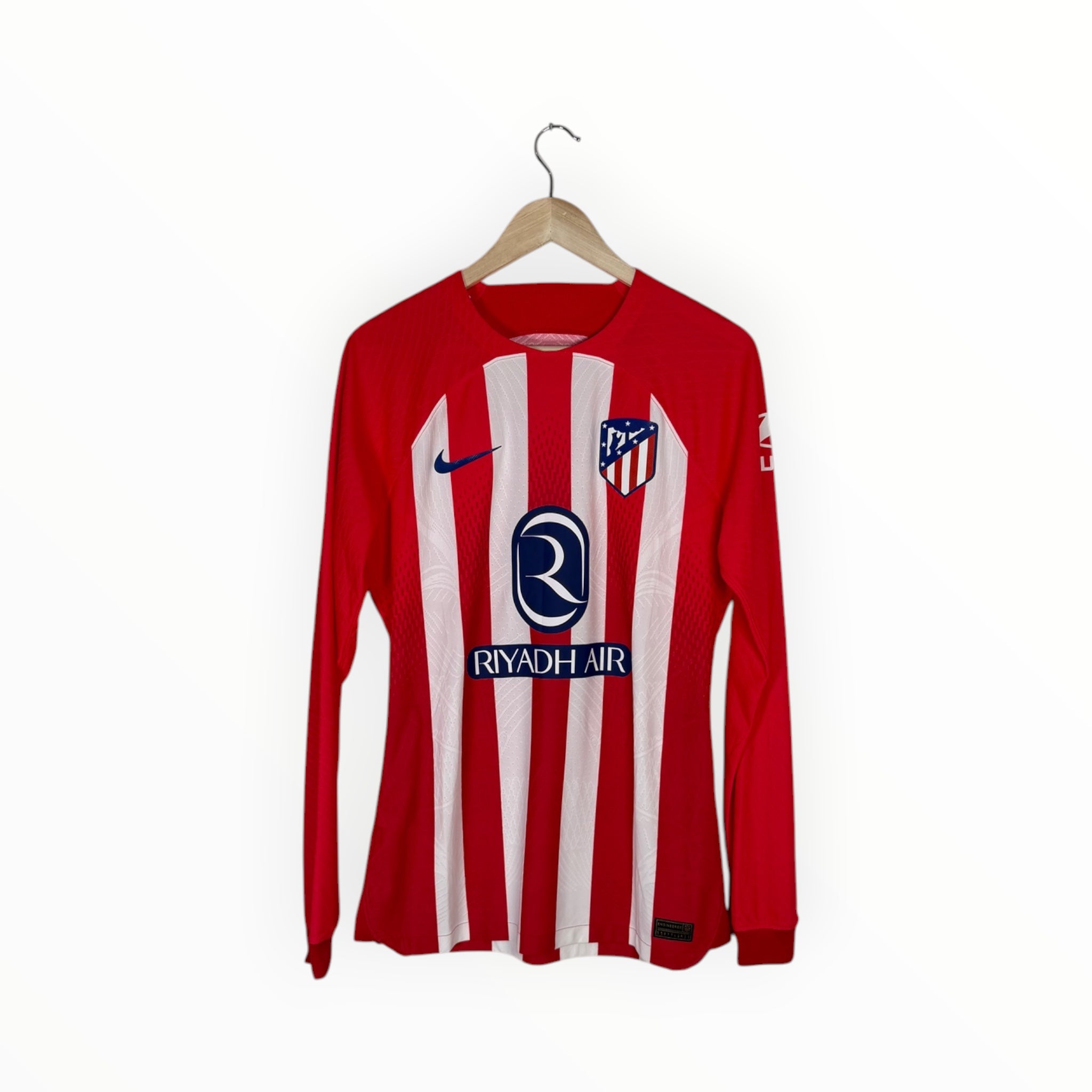 A red and white vertically striped long-sleeve Atletico Madrid soccer jersey hanging on a wooden hanger against a plain white background. The jersey features the Nike swoosh, the club crest, and the Riyadh Air sponsor logo.