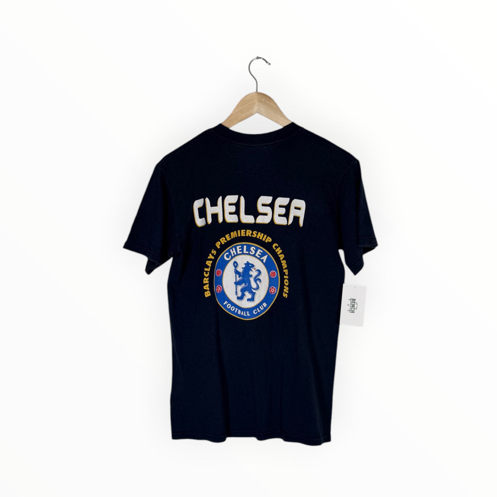 A navy blue Chelsea FC t-shirt hanging on a wooden hanger against a plain white wall. The back of the shirt features a large white 'CHELSEA' text graphic above a circular blue and white Chelsea crest.