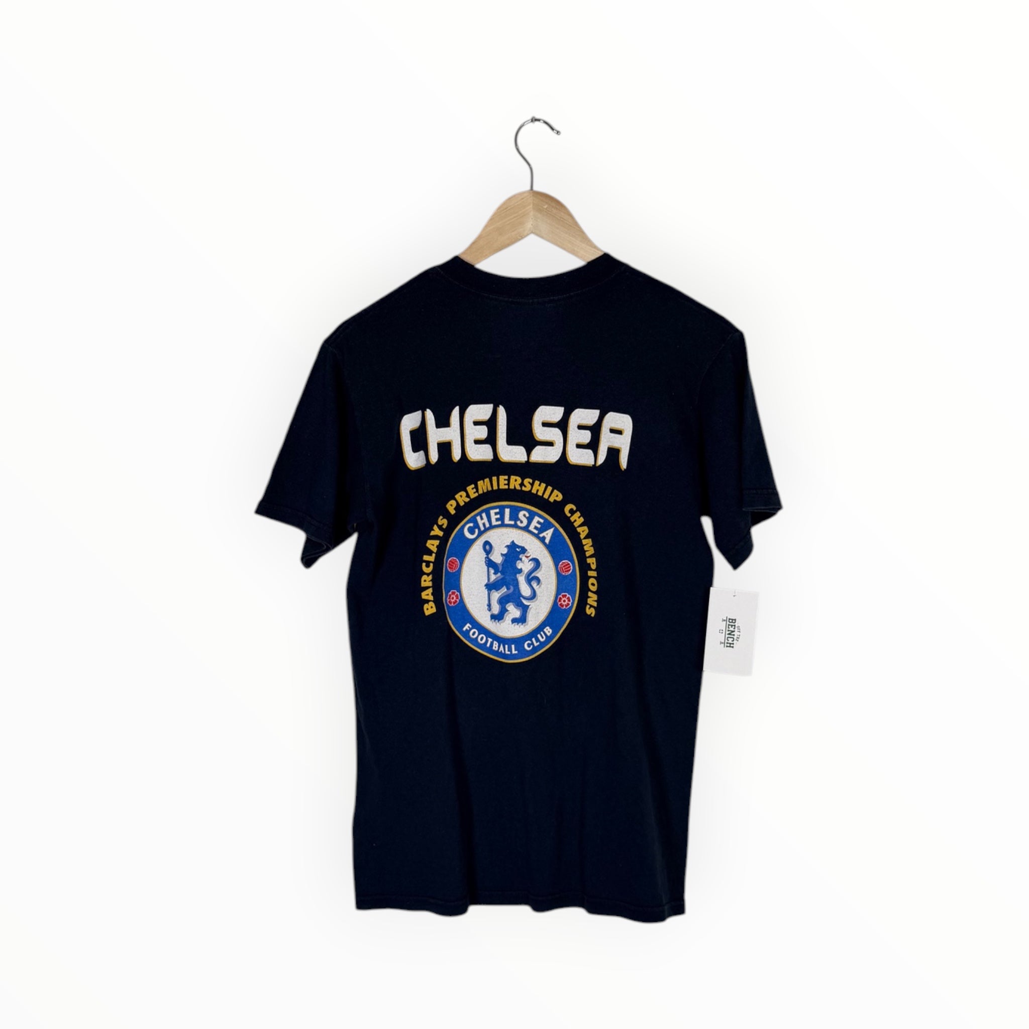 A navy blue Chelsea FC t-shirt hanging on a wooden hanger against a plain white wall. The back of the shirt features a large white 'CHELSEA' text graphic above a circular blue and white Chelsea crest.