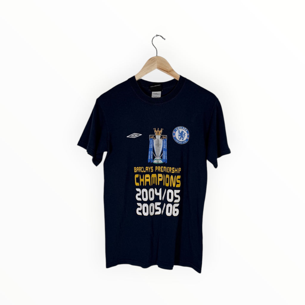 A navy blue Chelsea FC short-sleeve t-shirt hanging on a wooden hanger against a plain white background. The shirt features a graphic celebrating 2004/05 and 2005/06 Barclays Premiership Champions, with the Umbro logo on the right chest and the Chelsea crest on the left chest.