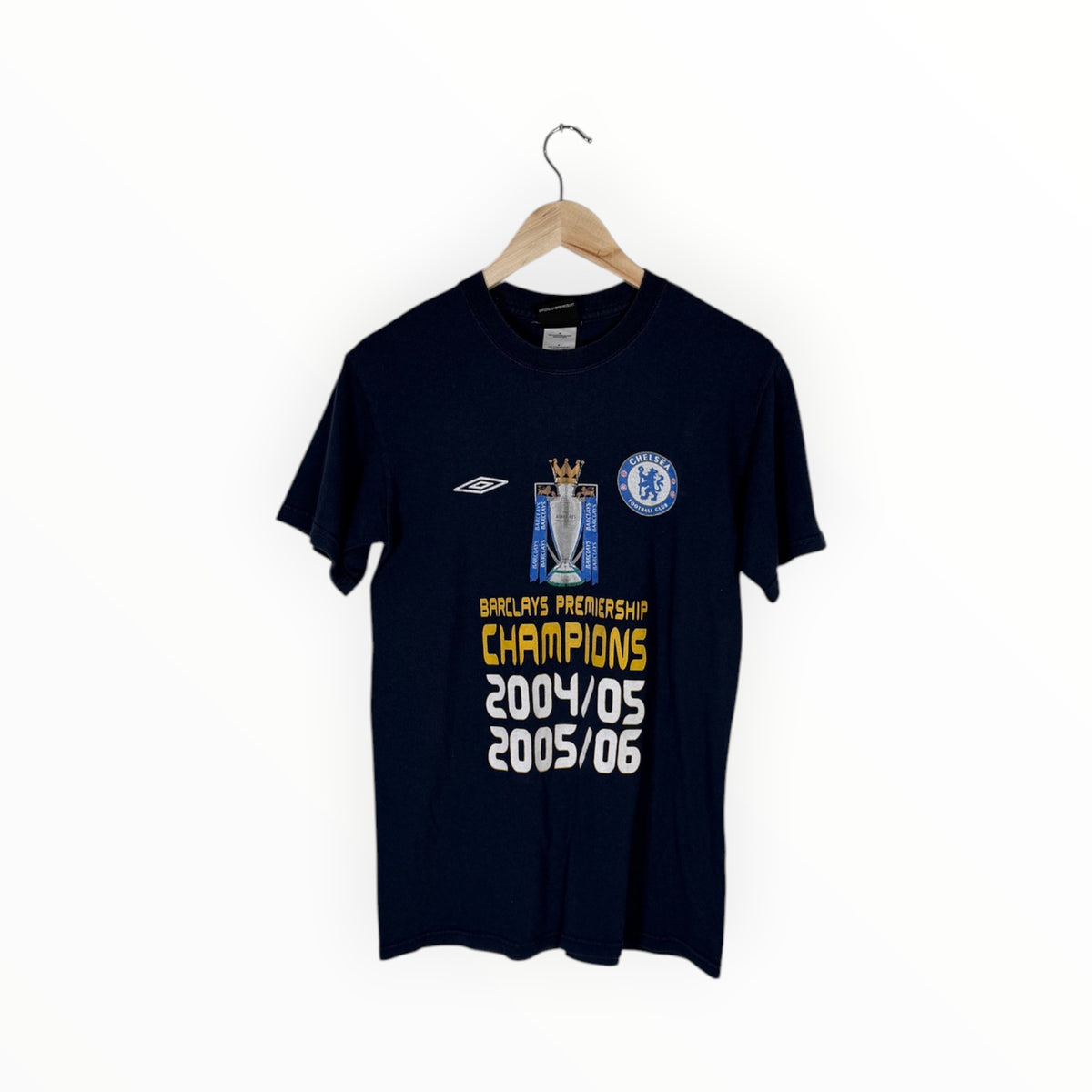 A navy blue Chelsea FC short-sleeve t-shirt hanging on a wooden hanger against a plain white background. The shirt features a graphic celebrating 2004/05 and 2005/06 Barclays Premiership Champions, with the Umbro logo on the right chest and the Chelsea crest on the left chest.