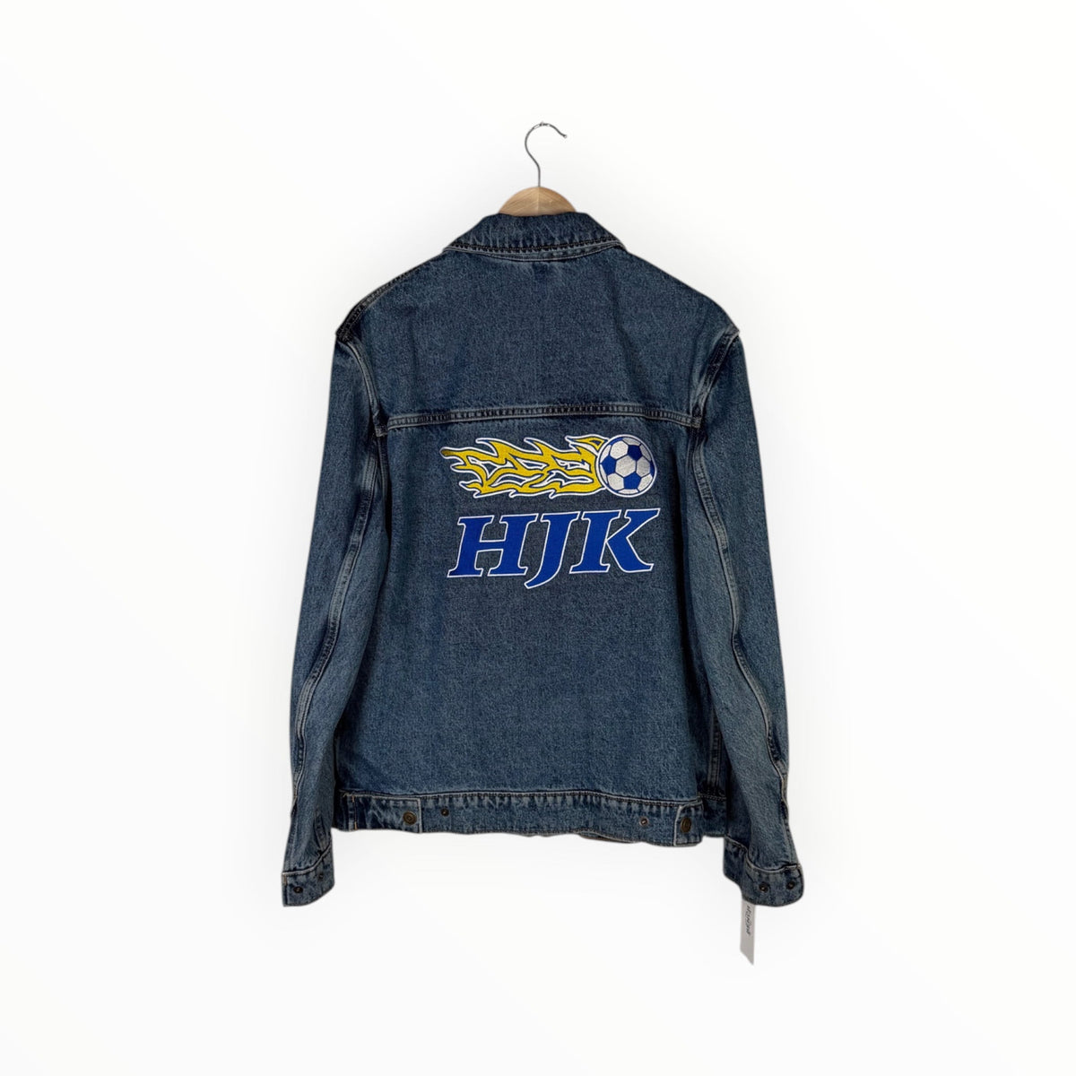 A blue denim jacket hanging on a wooden hanger against a white background, featuring a large graphic on the back with a yellow and blue flame, a soccer ball, and the text 'HJK' in blue.