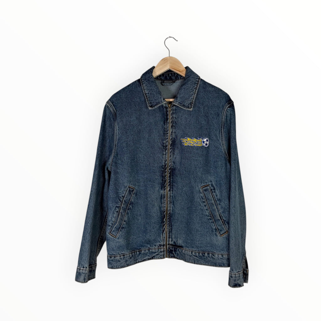 A dark blue denim jacket hanging on a wooden hanger against a plain white background. The jacket features a small embroidered graphic on the left chest with the text 'STOCKER' and a soccer ball.