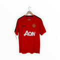 A red Manchester United soccer jersey hanging on a wooden hanger against a plain white background. The jersey features the white AON sponsor logo across the chest, a black Nike swoosh on the right chest, and the Manchester United club crest on the left chest.