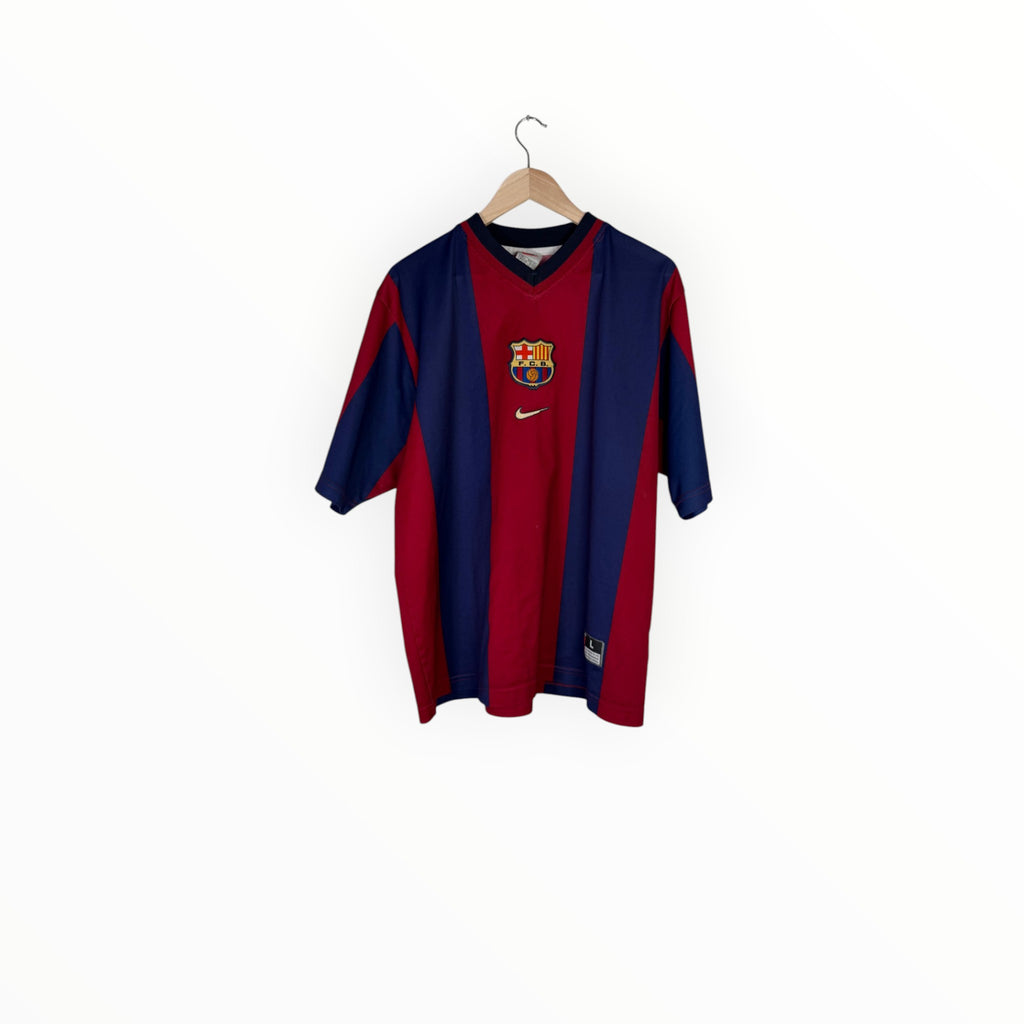 A vintage FC Barcelona home soccer jersey by Nike, featuring navy blue and red vertical stripes with the club crest on the chest, hanging on a wooden hanger against a plain white background.