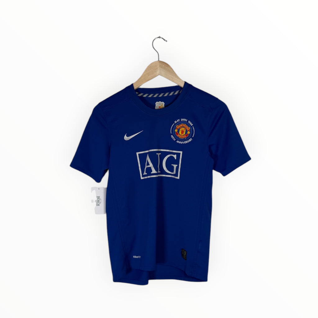 A blue Manchester United Nike soccer jersey hanging on a wooden hanger against a white background, featuring the AIG sponsor logo and Manchester United crest.