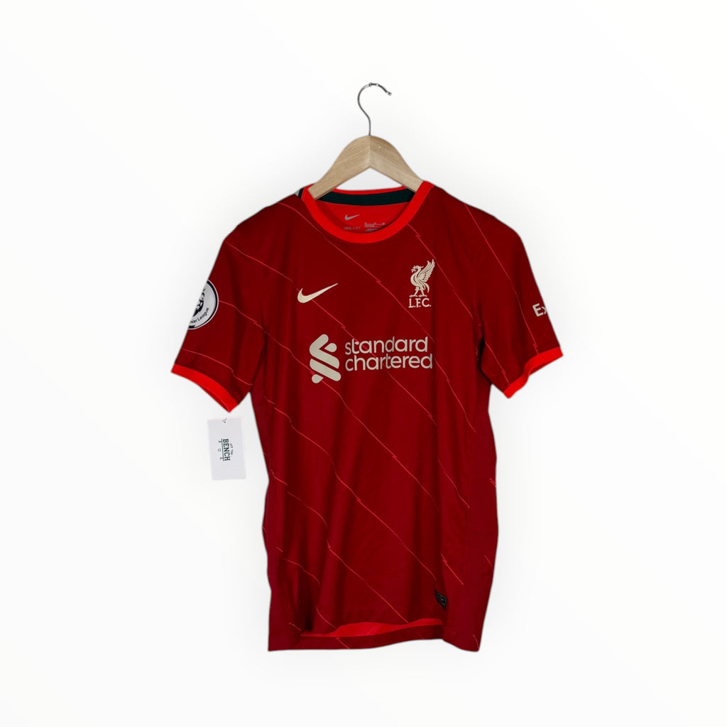 A red Liverpool football club home jersey with white logos and subtle diagonal stripe patterns, hanging on a wooden hanger against a plain white background.