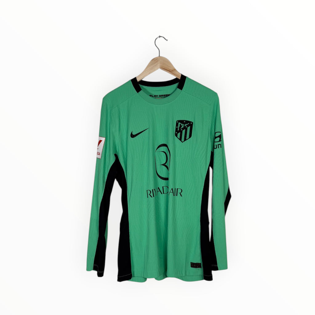 A green long-sleeve athletic jersey with black side panels and black logos, hanging on a wooden hanger against a plain white background. Visible branding includes a Nike swoosh, an Atlético Madrid crest, and a sponsor logo reading Rivalair.