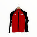 A red and black Puma track jacket with white shoulder stripes, hanging on a wooden hanger against a plain white background.