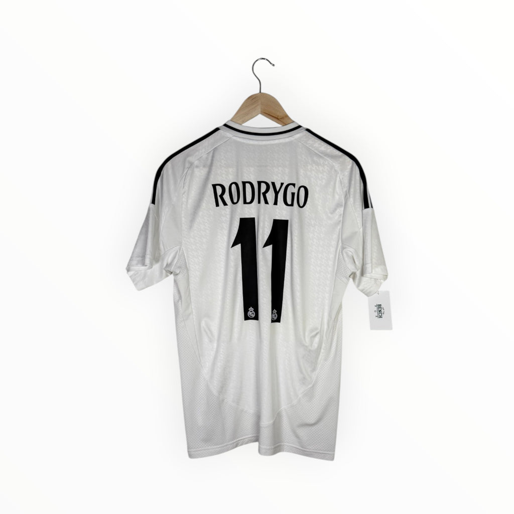 A white Real Madrid jersey with 'RODRYGO' and the number '11' printed in black on the back, hanging on a wooden hanger against a white background.