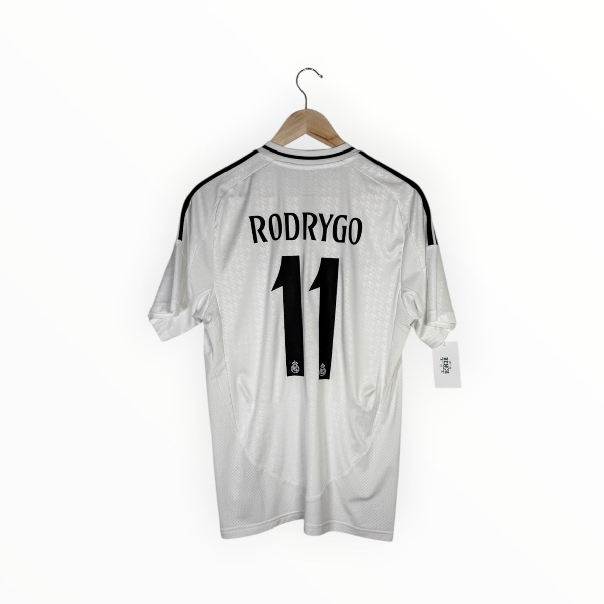 A white Real Madrid jersey with 'RODRYGO' and the number '11' printed in black on the back, hanging on a wooden hanger against a white background.