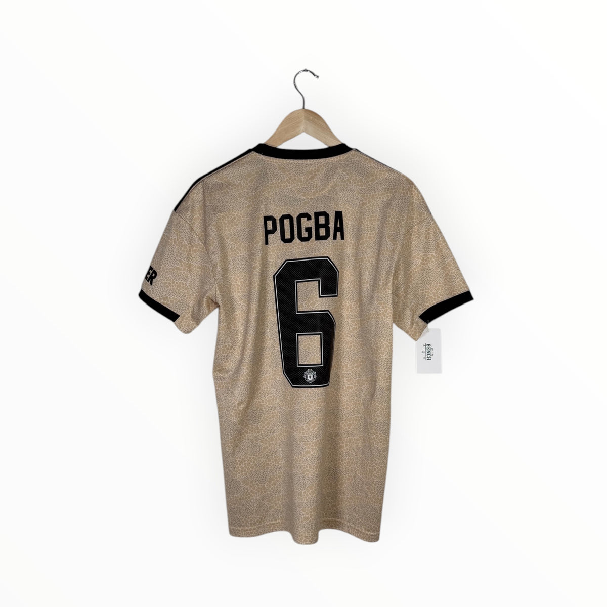 A beige soccer jersey with black trim, featuring the name 'POGBA' and number '6' on the back, hanging on a wooden hanger against a white background.