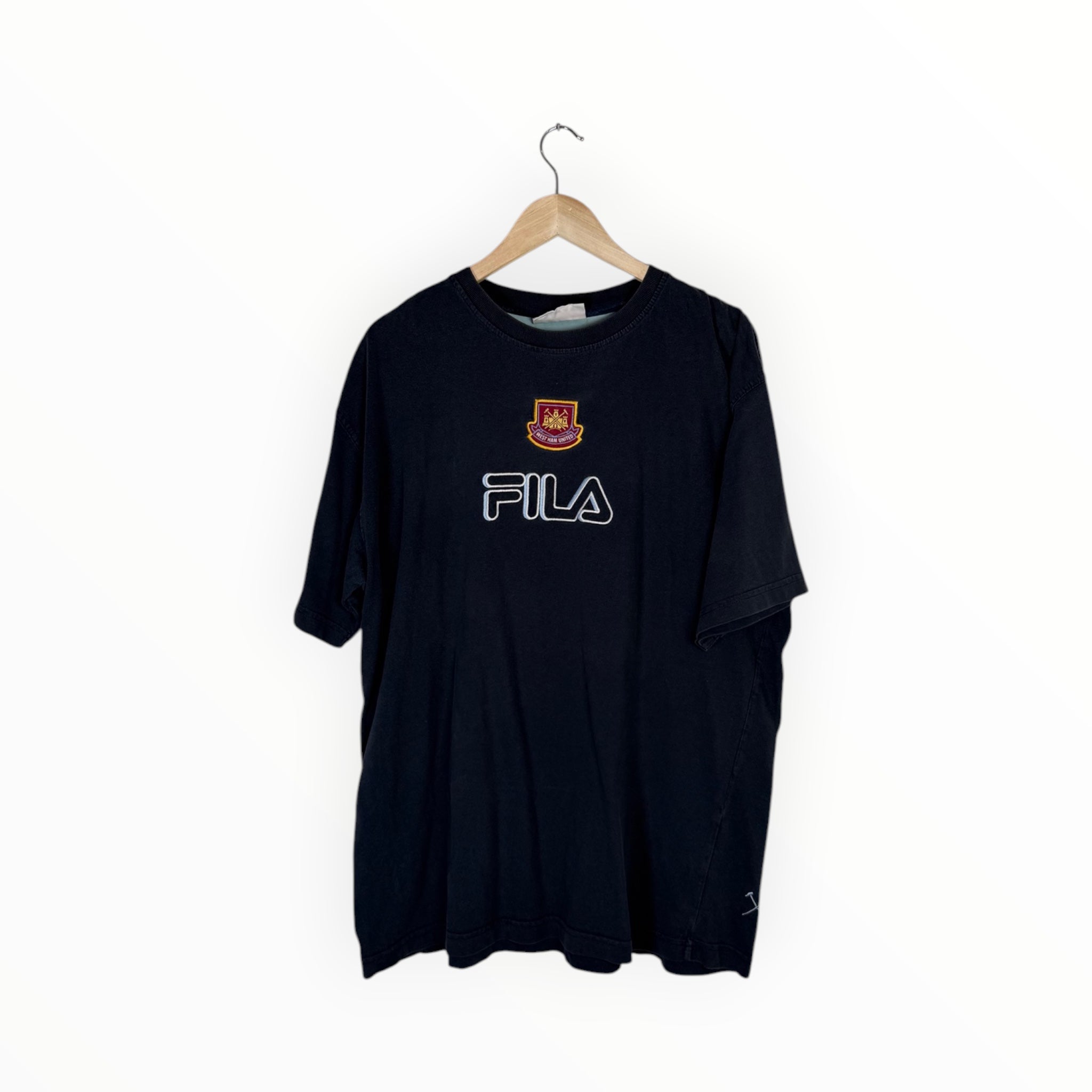 A black FILA t-shirt hanging on a wooden hanger against a plain white background. The shirt features the FILA logo in white outlined lettering across the chest and a small red and gold club crest above it.